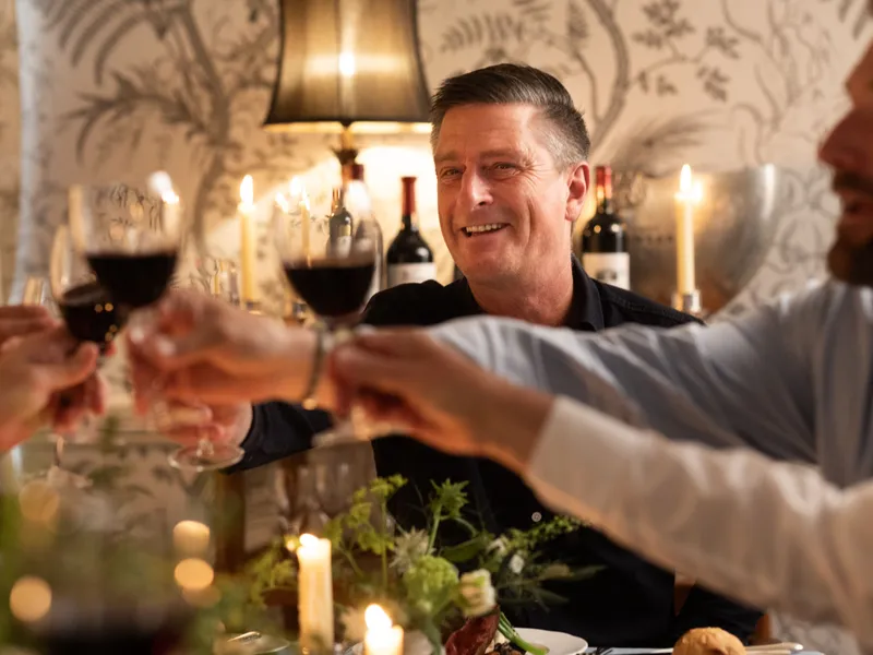 Friends raising glasses of red wine in a candlelit restaurant while a smiling man sits at the centre of the table.
