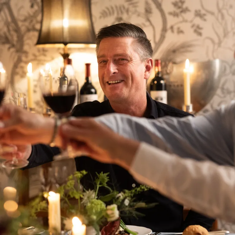 Friends raising glasses of red wine in a candlelit restaurant while a smiling man sits at the centre of the table.