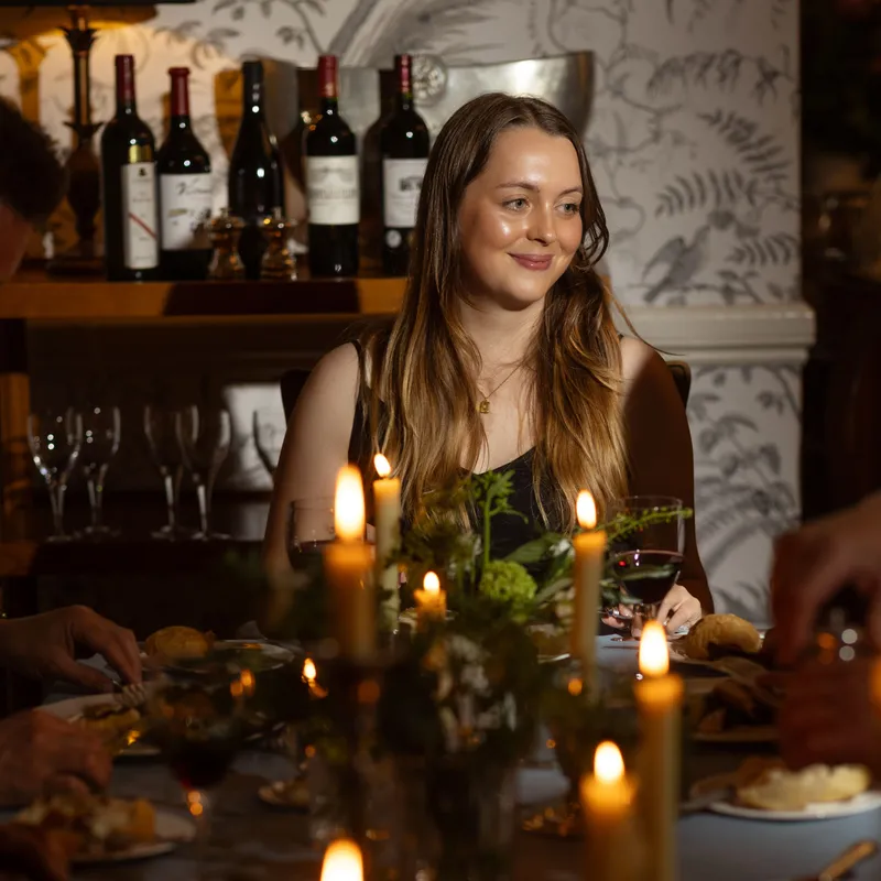 Woman seated at a candlelit dinner table with wine bottles in the background