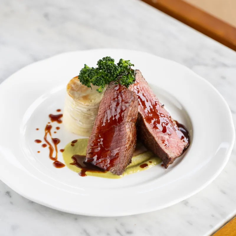 Plated sliced duck breast with glossy sauce, fondant potato and tenderstem broccoli on a white plate