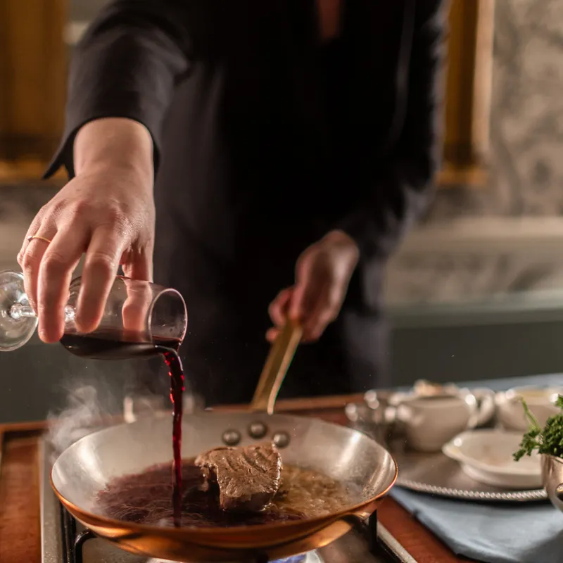 Chef pouring red wine into a frying pan with steak on a hob