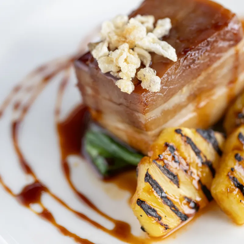 Close-up of glazed pork belly topped with crispy crackling, served with green vegetables, grilled pineapple and a rich sauce.