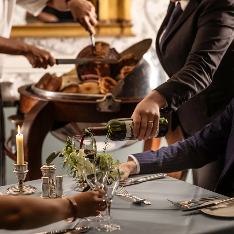 Sommelier pouring red wine for a guest at an elegant restaurant table while chefs carve roast meat in the background