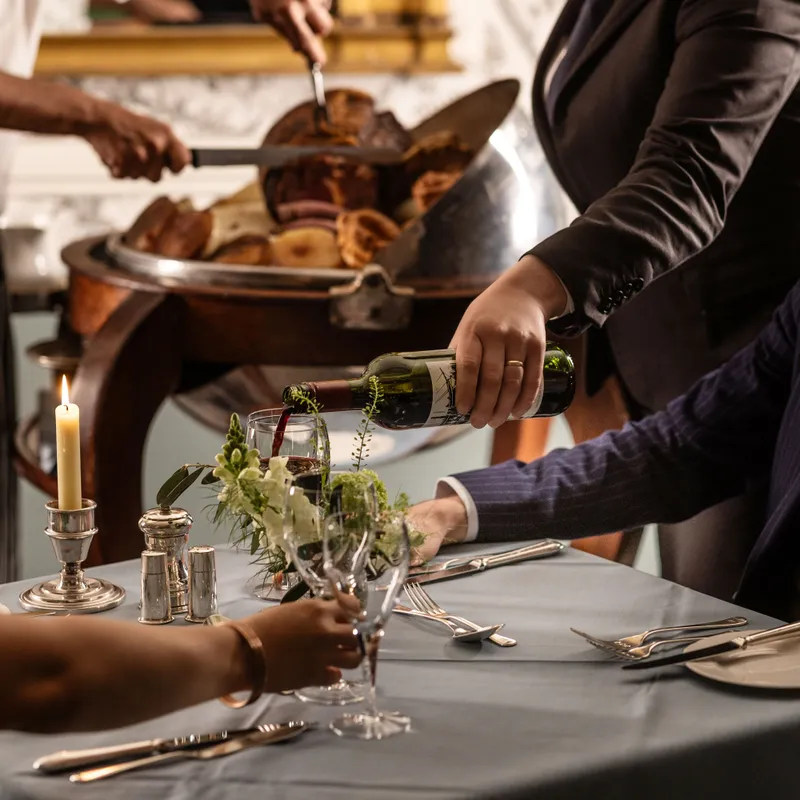 Sommelier pouring red wine for a guest at an elegant restaurant table while chefs carve roast meat in the background