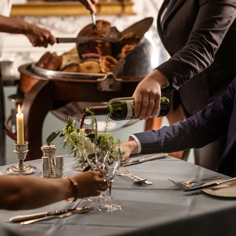 Sommelier pouring red wine for a guest at an elegant restaurant table while chefs carve roast meat in the background
