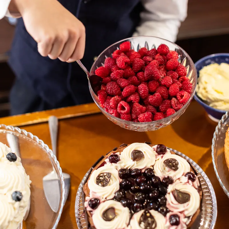 Person in a dark waistcoat serving fresh raspberries beside decorated cakes on a wooden table