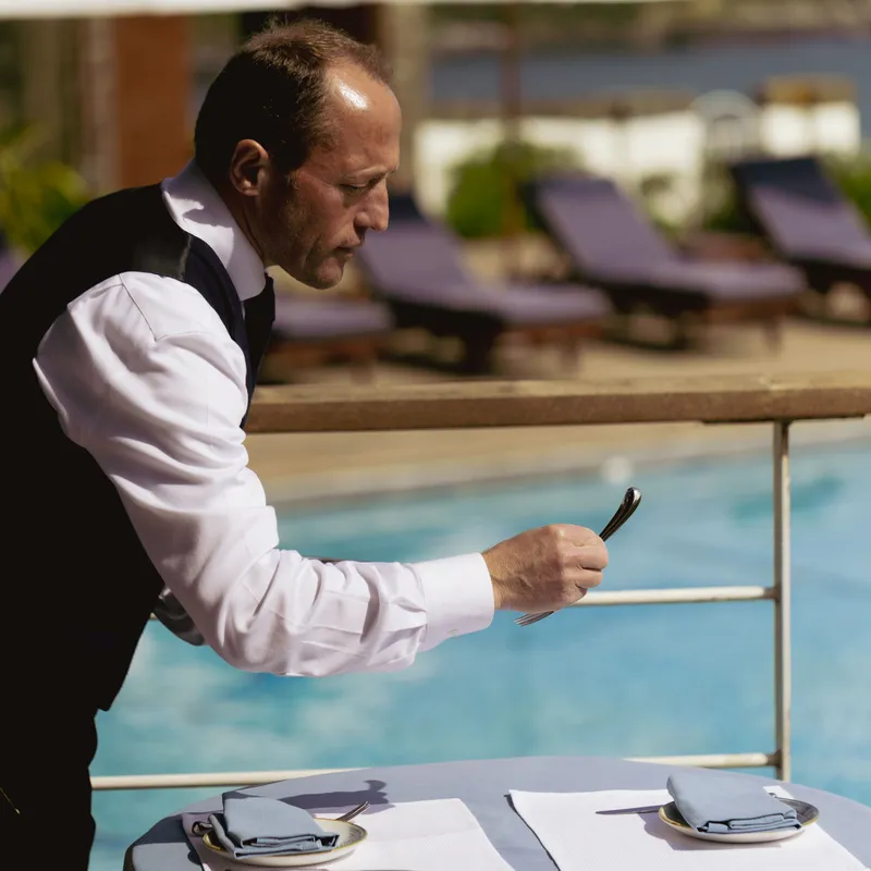 Waiter arranging cutlery on a poolside dining table at an outdoor restaurant