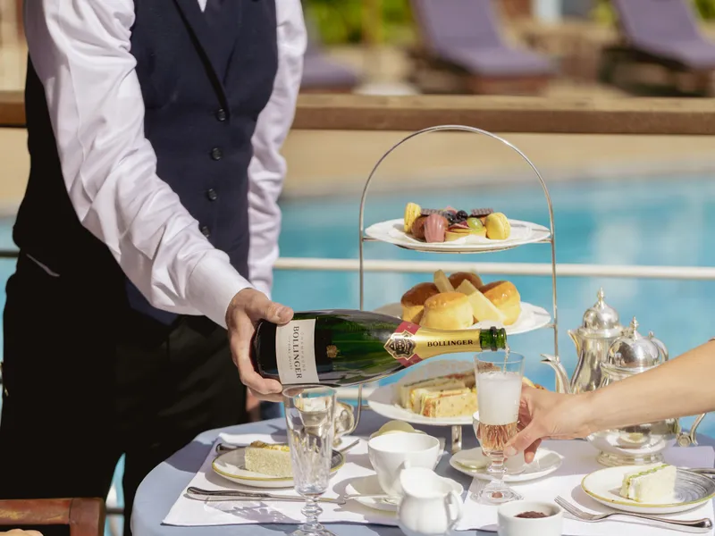 Waiter pouring champagne into a glass beside a poolside afternoon tea table