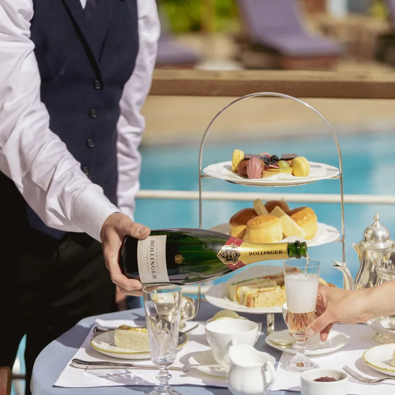 Waiter pouring champagne into a glass beside a poolside afternoon tea table