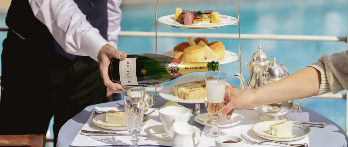 Waiter pouring champagne into a glass beside a poolside afternoon tea table