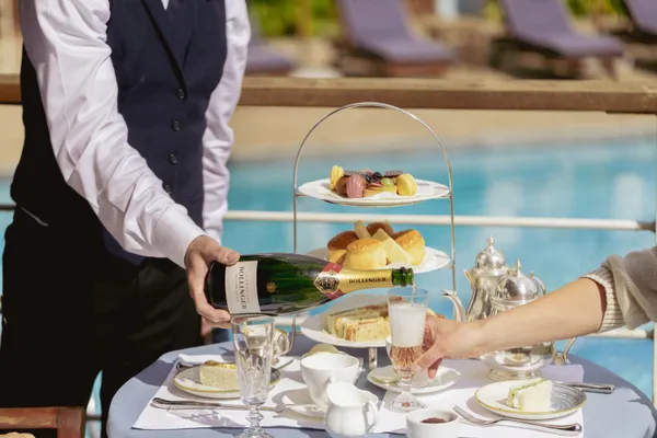 Waiter pouring champagne into a glass beside a poolside afternoon tea table