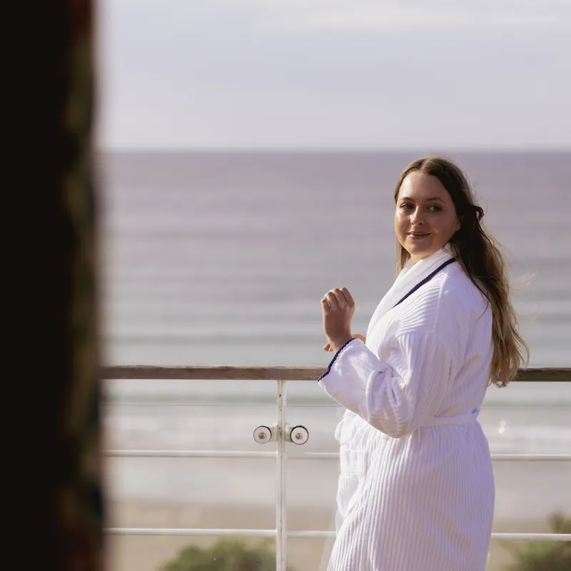 Woman in a white robe standing on a seaside balcony, looking back towards the camera