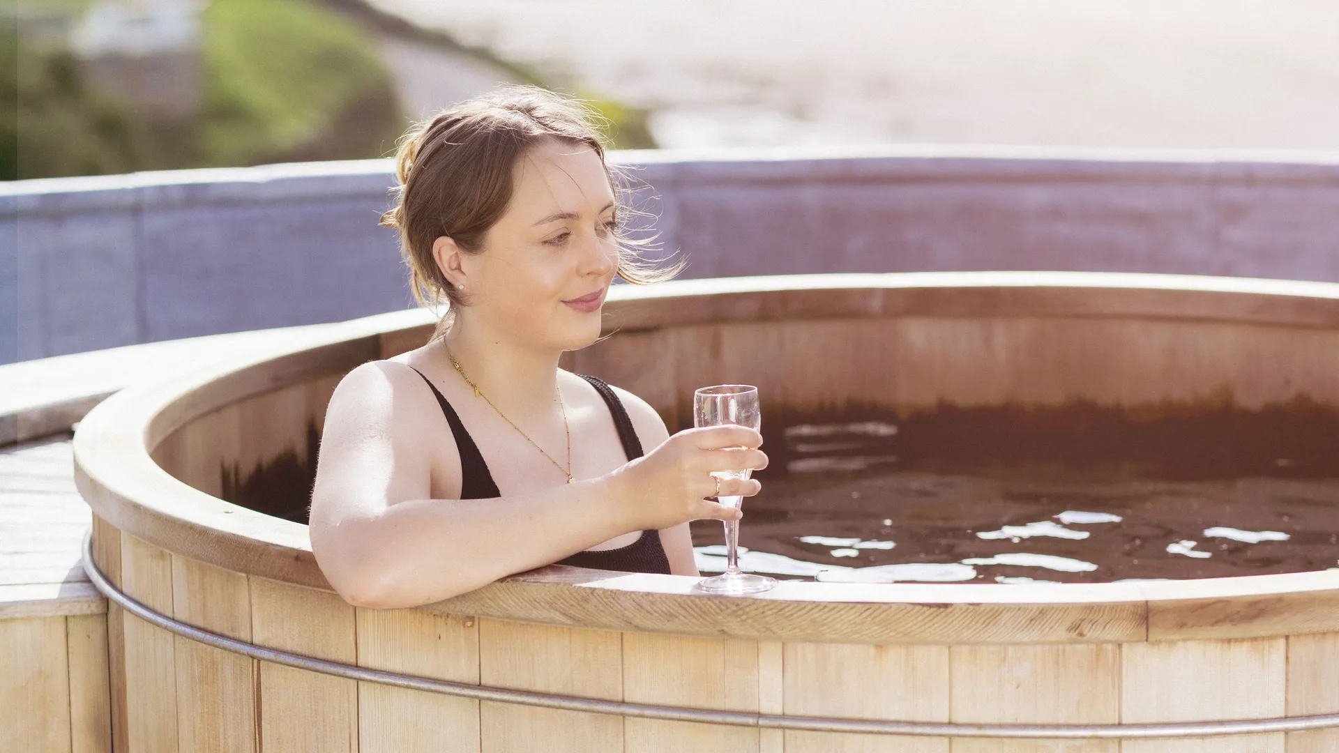 Woman relaxing in a wooden outdoor hot tub with a glass of sparkling wine beside a scenic river view