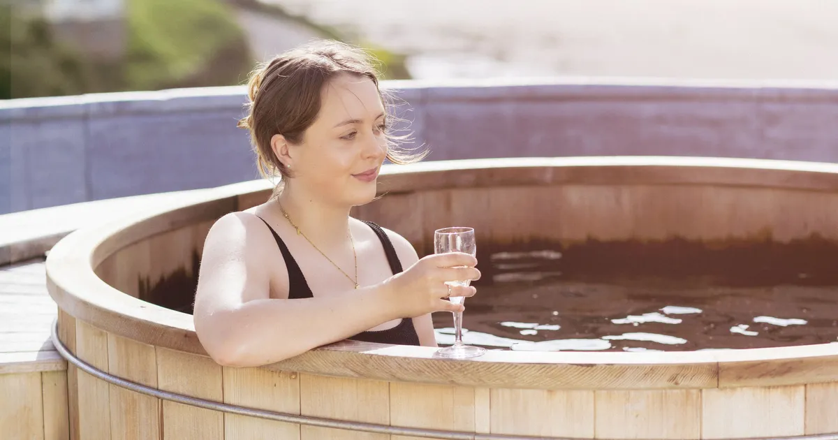 Woman relaxing in a wooden outdoor hot tub with a glass of sparkling wine beside a scenic river view