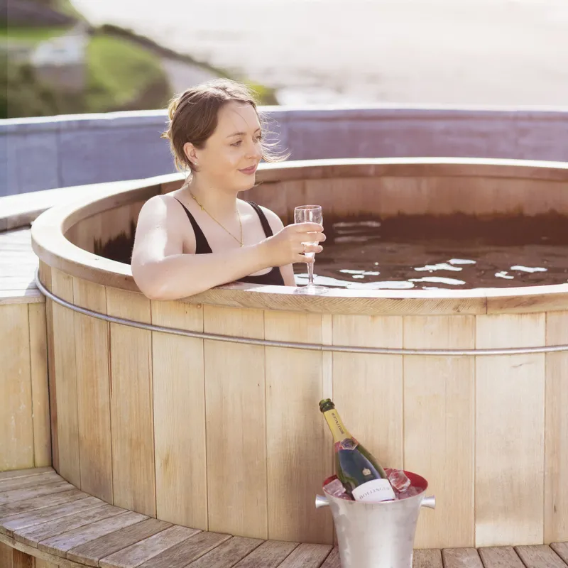 Woman relaxing in a wooden outdoor hot tub with a glass of sparkling wine beside a scenic river view