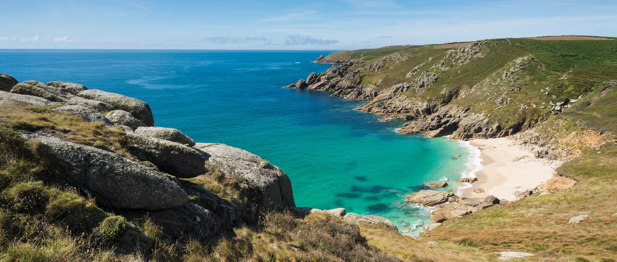 Rocky coastal cove with turquoise sea, sandy beach and grassy cliffs under a clear blue sky