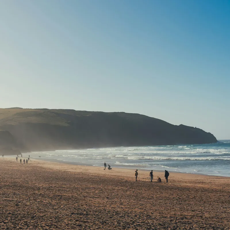 Wide sandy beach with people walking along the shoreline beneath rugged cliffs and a clear blue sky