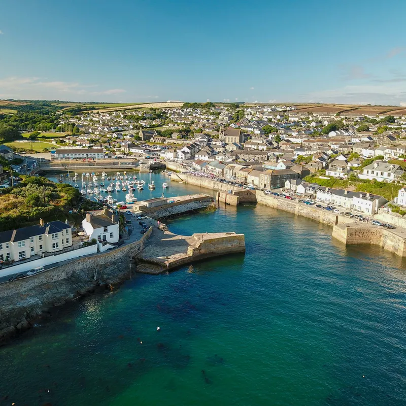 Aerial view of a coastal harbour town with turquoise water, stone sea walls, moored boats and tightly packed houses under a clear blue sky.