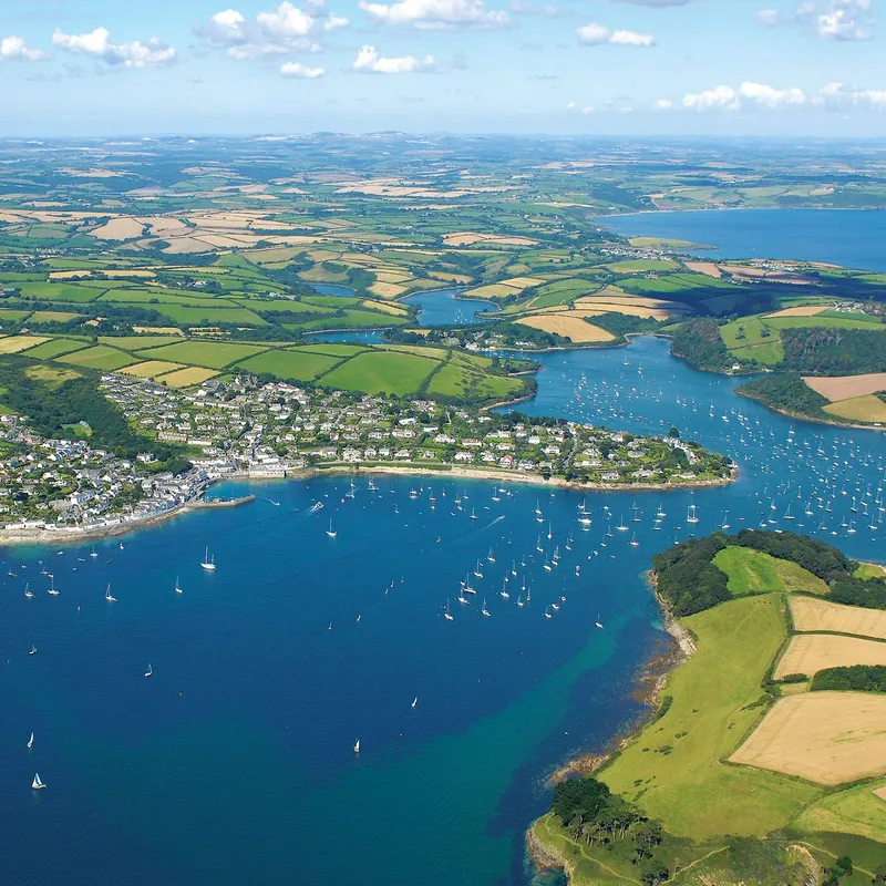 Aerial view of a coastal harbour with sailing boats, a waterside village and patchwork fields stretching inland under a blue sky.