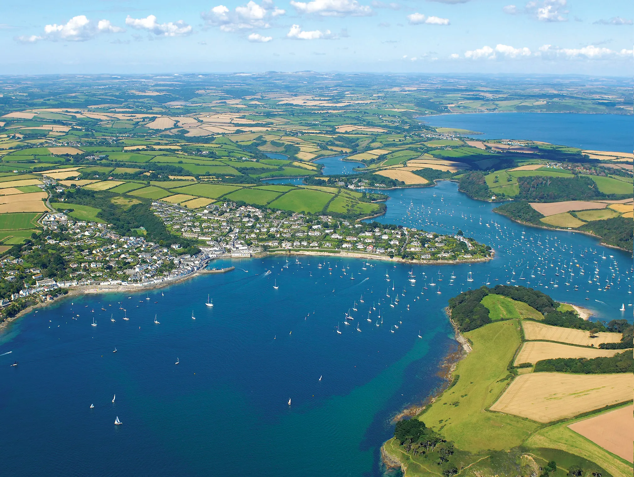 Aerial view of a coastal harbour with sailing boats, a waterside village and patchwork fields stretching inland under a blue sky.