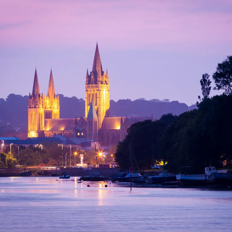 Illuminated cathedral with tall spires beside a river at dusk under a pink and purple sky