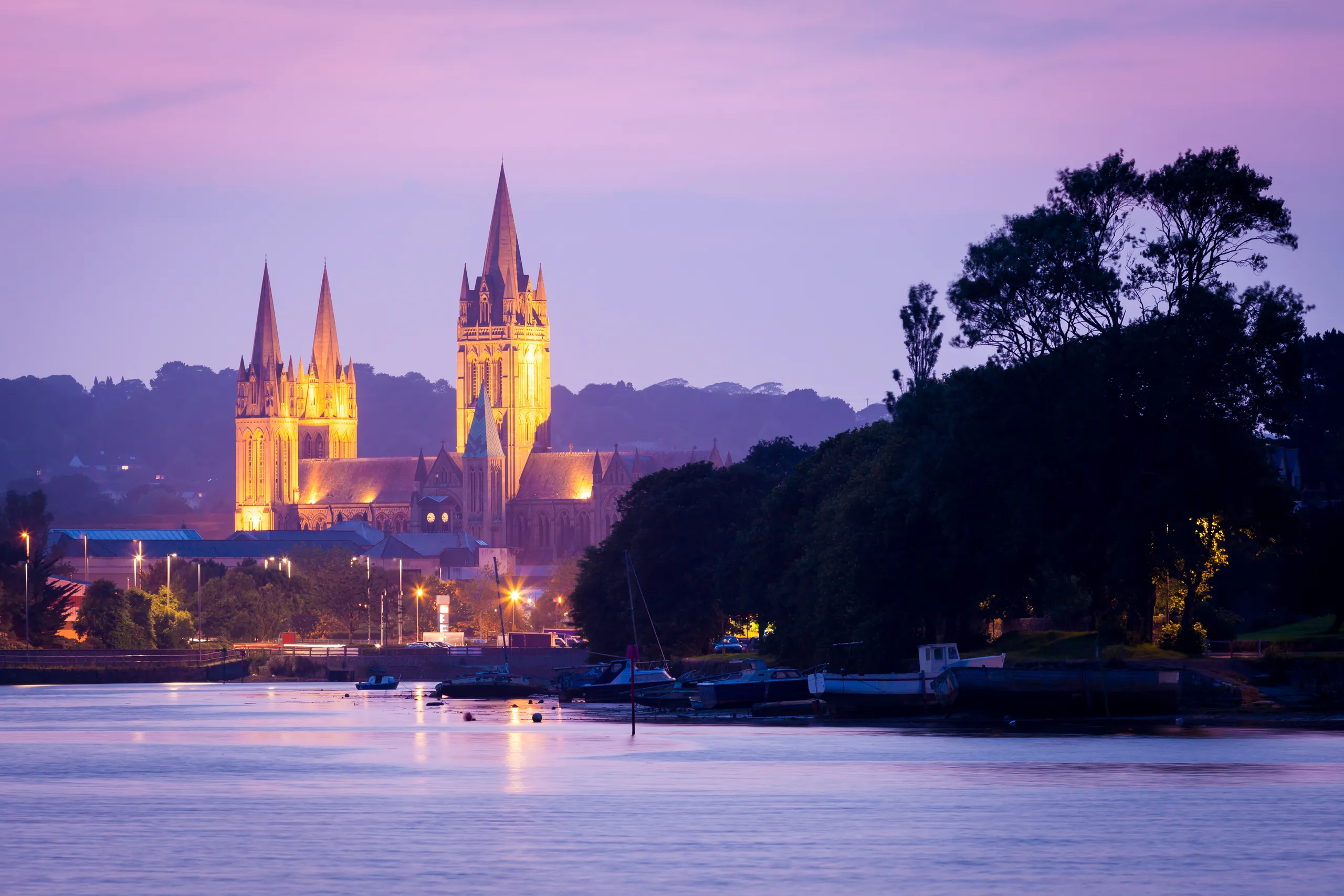 Illuminated cathedral with tall spires beside a river at dusk under a pink and purple sky