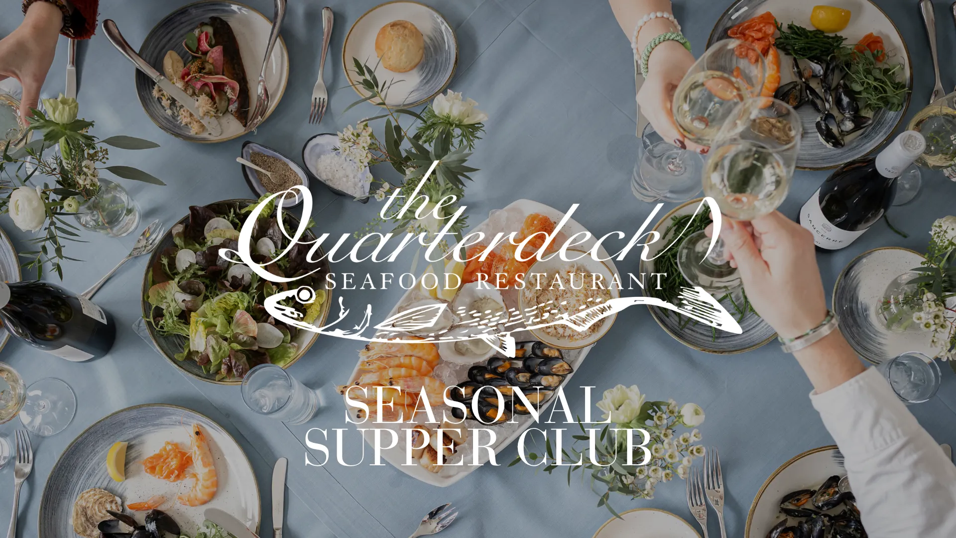 Overhead view of a seafood dining table with platters, salads, wine glasses and guests toasting, featuring The Quarterdeck Seafood Restaurant Seasonal Supper Club text.