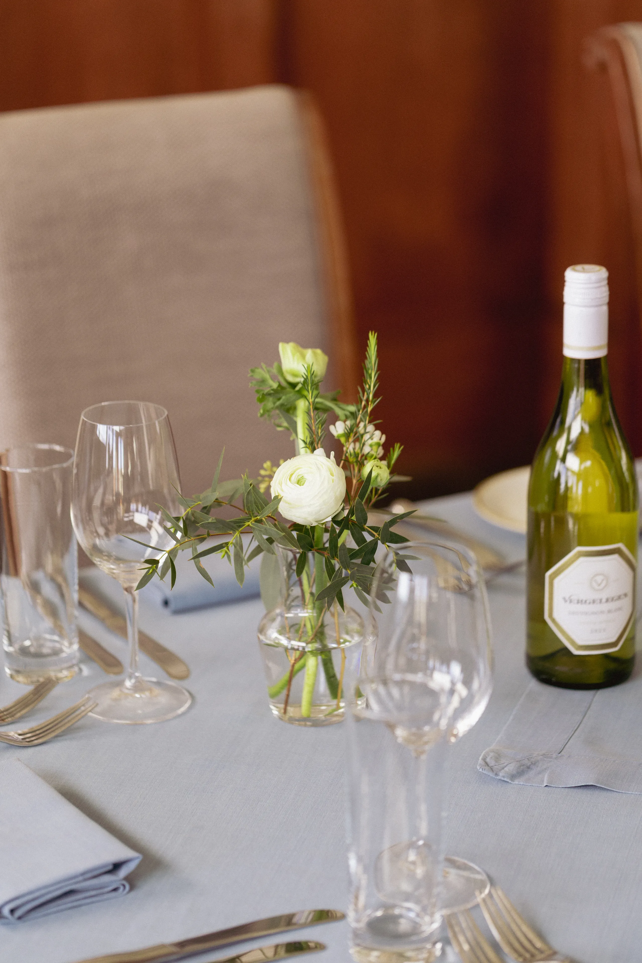 Elegantly set dining table with wine glasses, a bottle of white wine, and a small floral arrangement in a glass vase