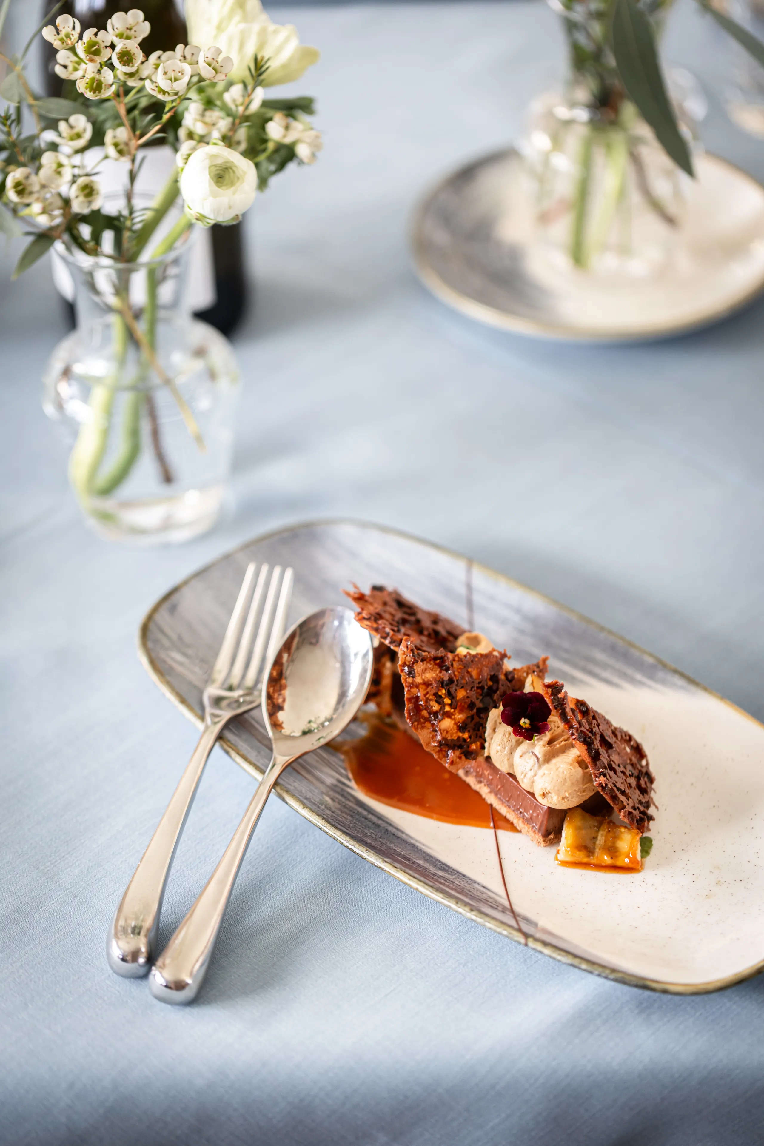 Elegant plated chocolate dessert with cream and caramel on a softly lit restaurant table with flowers in glass vases
