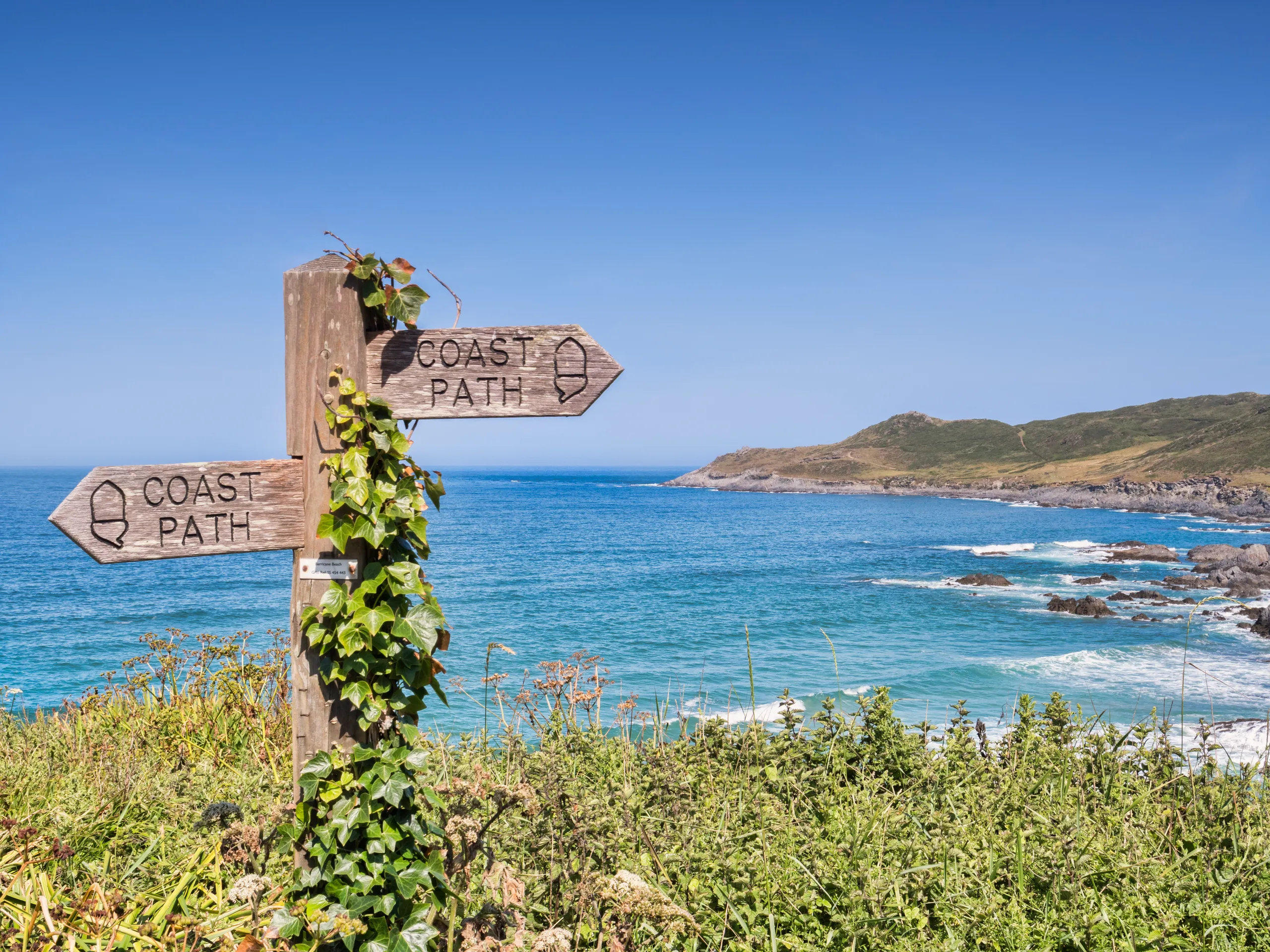South West Coast Path Wooden coast path signpost beside a turquoise sea and rocky headland under a clear blue sky
