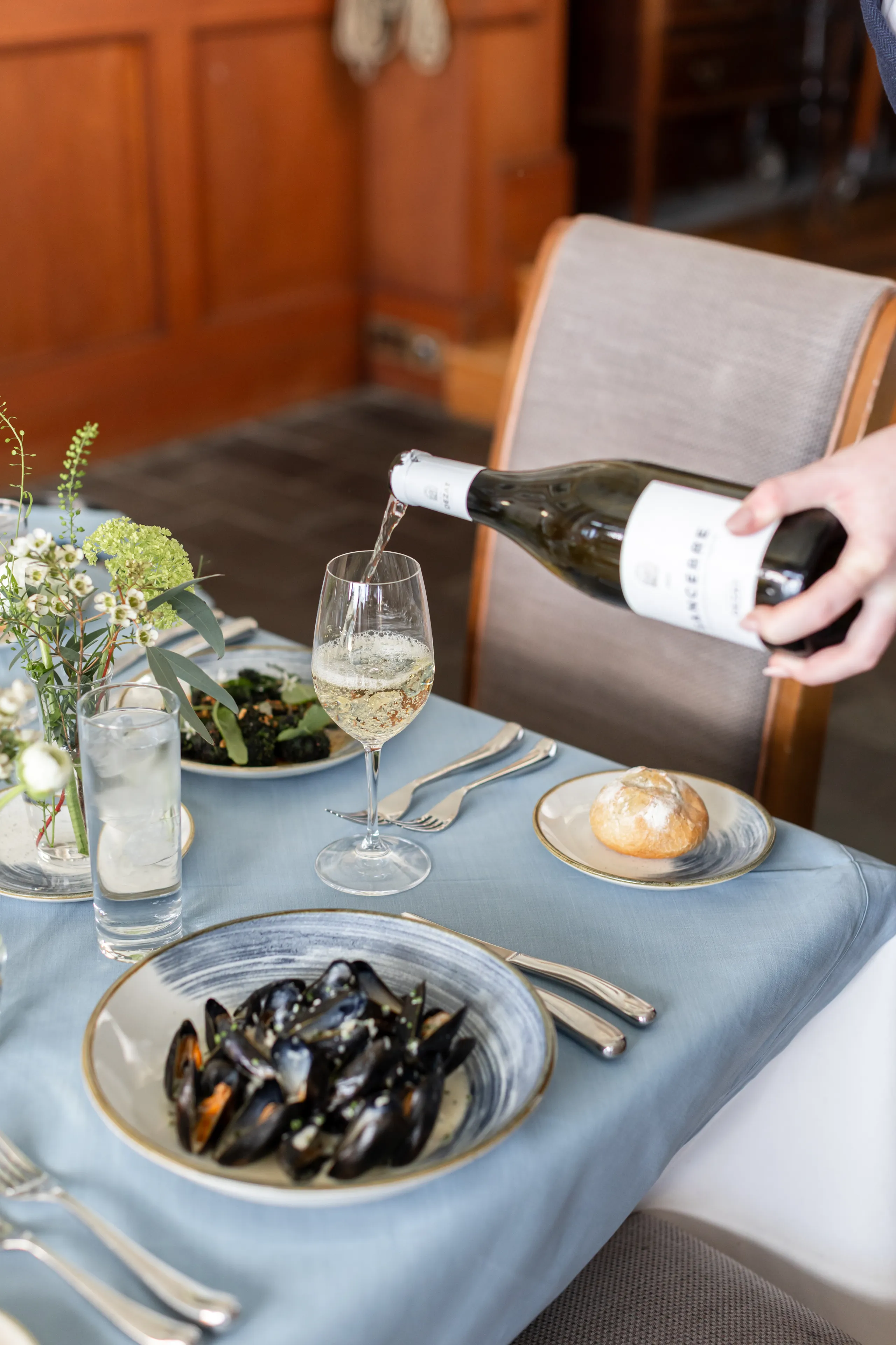 A bottle of white wine being poured into a glass at an elegantly set dining table with mussels, bread and floral arrangements.