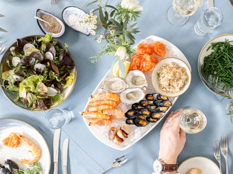 Overhead view of a seafood platter with oysters, prawns, mussels, smoked salmon and crab, served with salad and samphire on a light blue table.