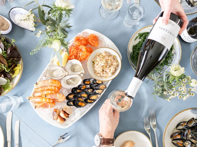 Overhead view of a seafood feast on a pale blue tablecloth, with oysters, prawns, mussels, salmon and crab alongside salad, greens and white wine being poured.