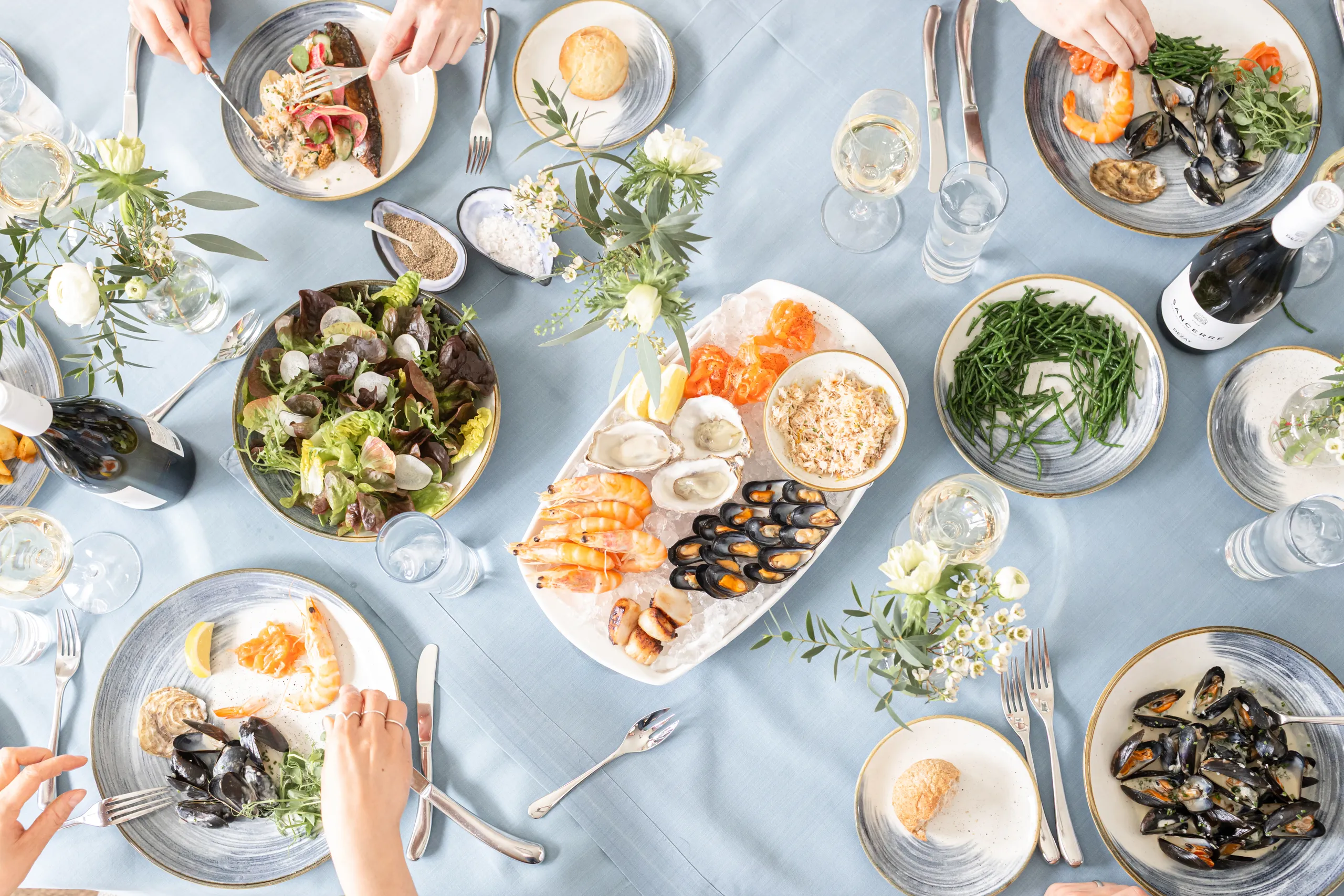 Overhead view of a seafood dining table with prawns, oysters, mussels, salad, green beans, wine and place settings on a pale blue tablecloth.