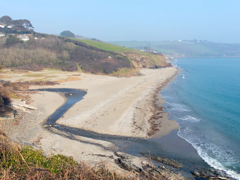 Gerrans Bay Wide view of a quiet sandy beach and turquoise sea below rugged cliffs and rolling countryside.