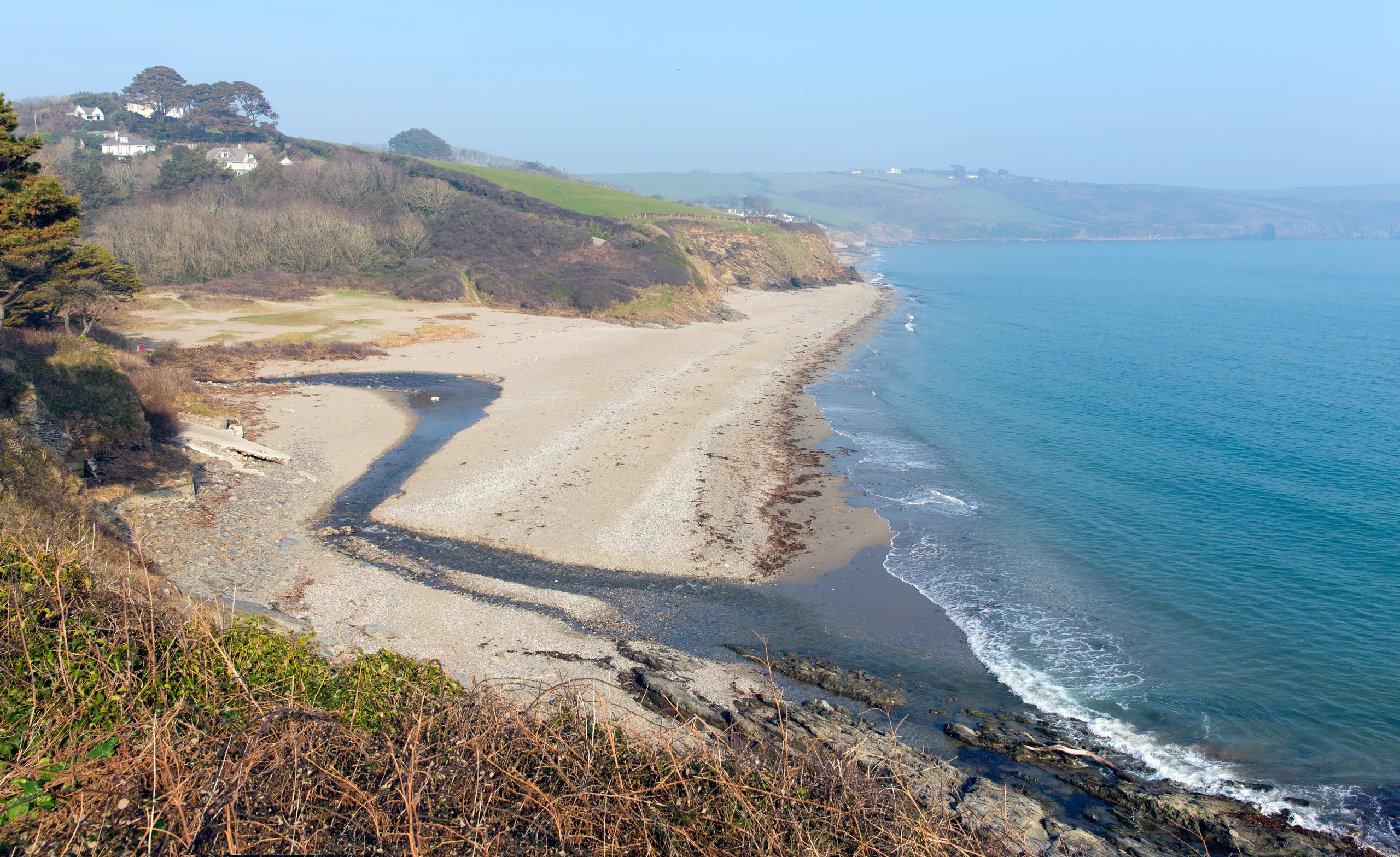 Gerrans Bay Wide view of a quiet sandy beach and turquoise sea below rugged cliffs and rolling countryside.