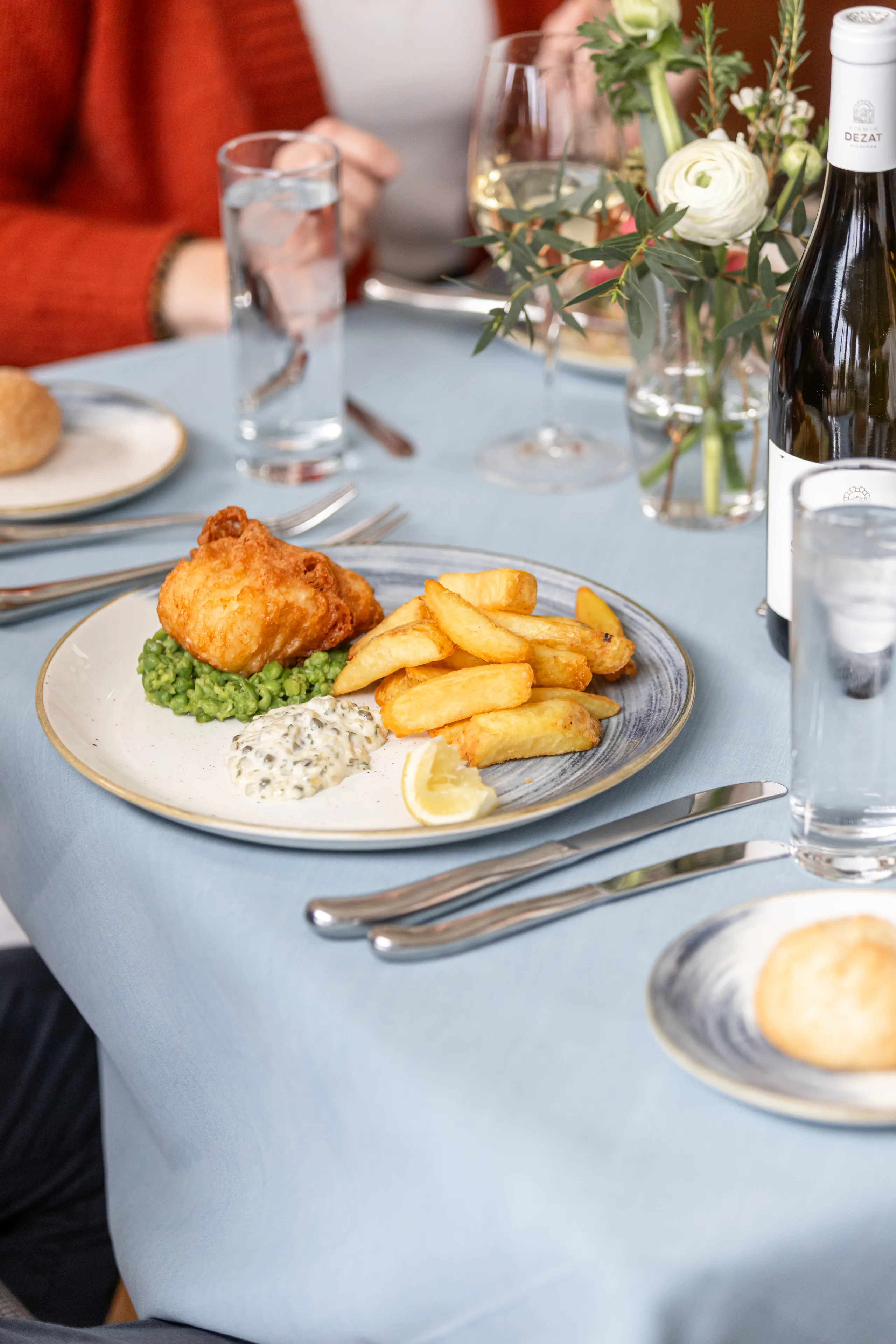 Plate of battered fish with chunky chips, mushy peas, tartare sauce and a lemon wedge on a restaurant table set with wine, water and flowers.