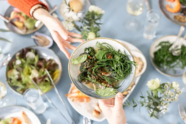 Overhead view of hands passing a bowl of green vegetables over a shared dining table with salads, seafood and glasses