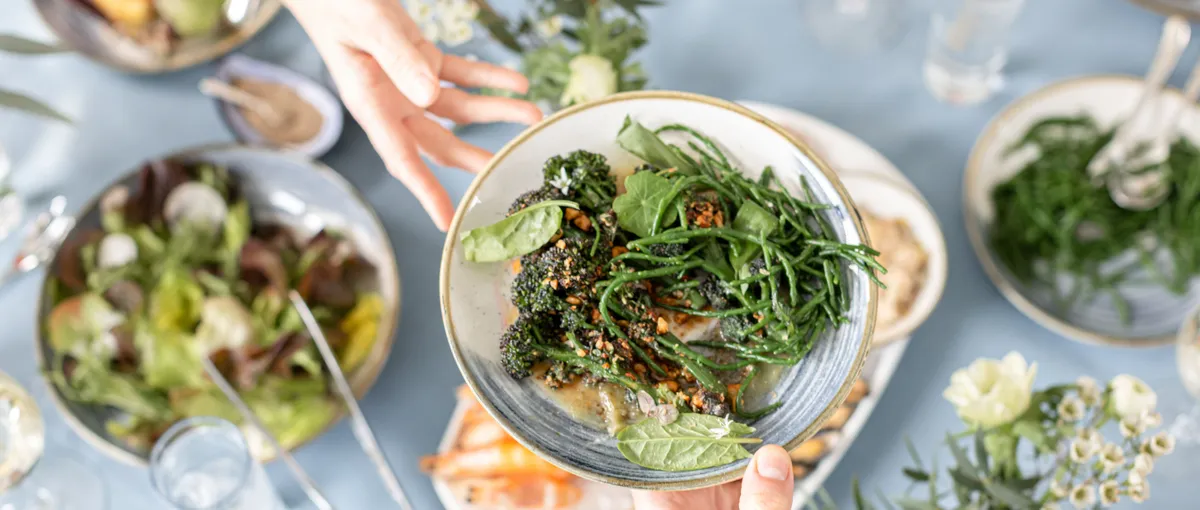 Overhead view of hands passing a bowl of green vegetables over a shared dining table with salads, seafood and glasses