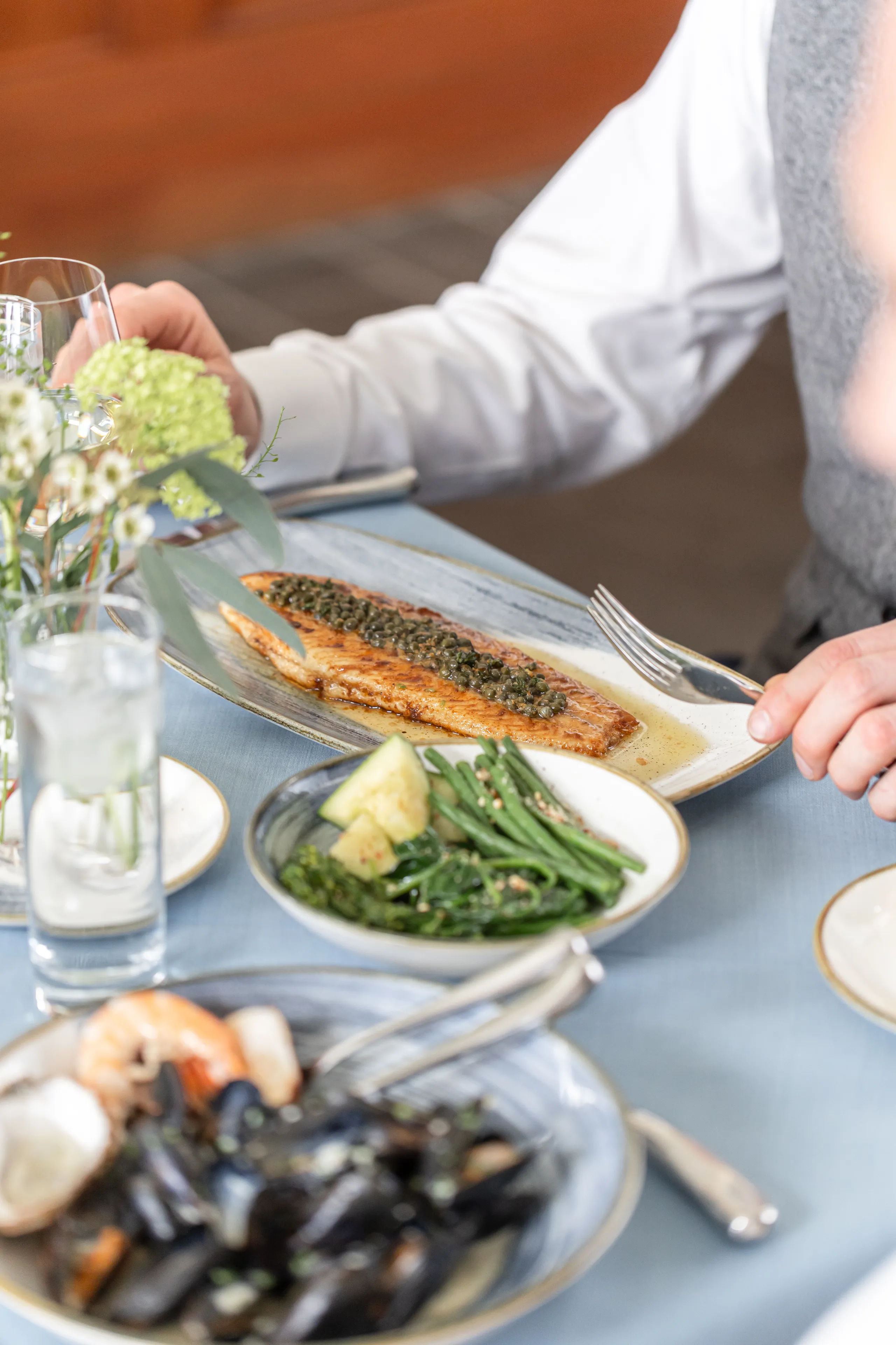 Diner at a restaurant table with a pan-fried fish fillet topped with capers, served alongside green vegetables and seafood dishes