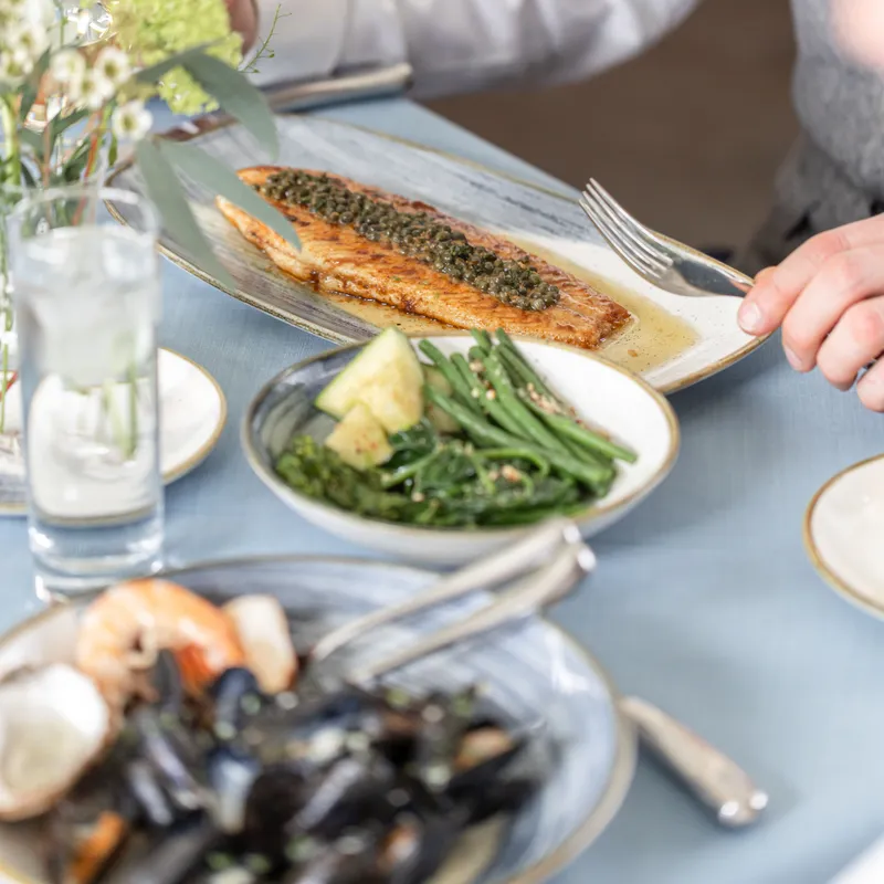 Diner at a restaurant table with a pan-fried fish fillet topped with capers, served alongside green vegetables and seafood dishes