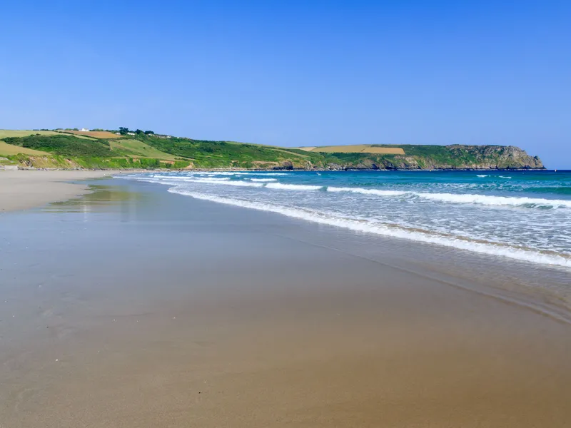 Pendower Beach Cornwall  Wide sandy beach with gentle waves, green hills and a rocky headland beneath a clear blue sky