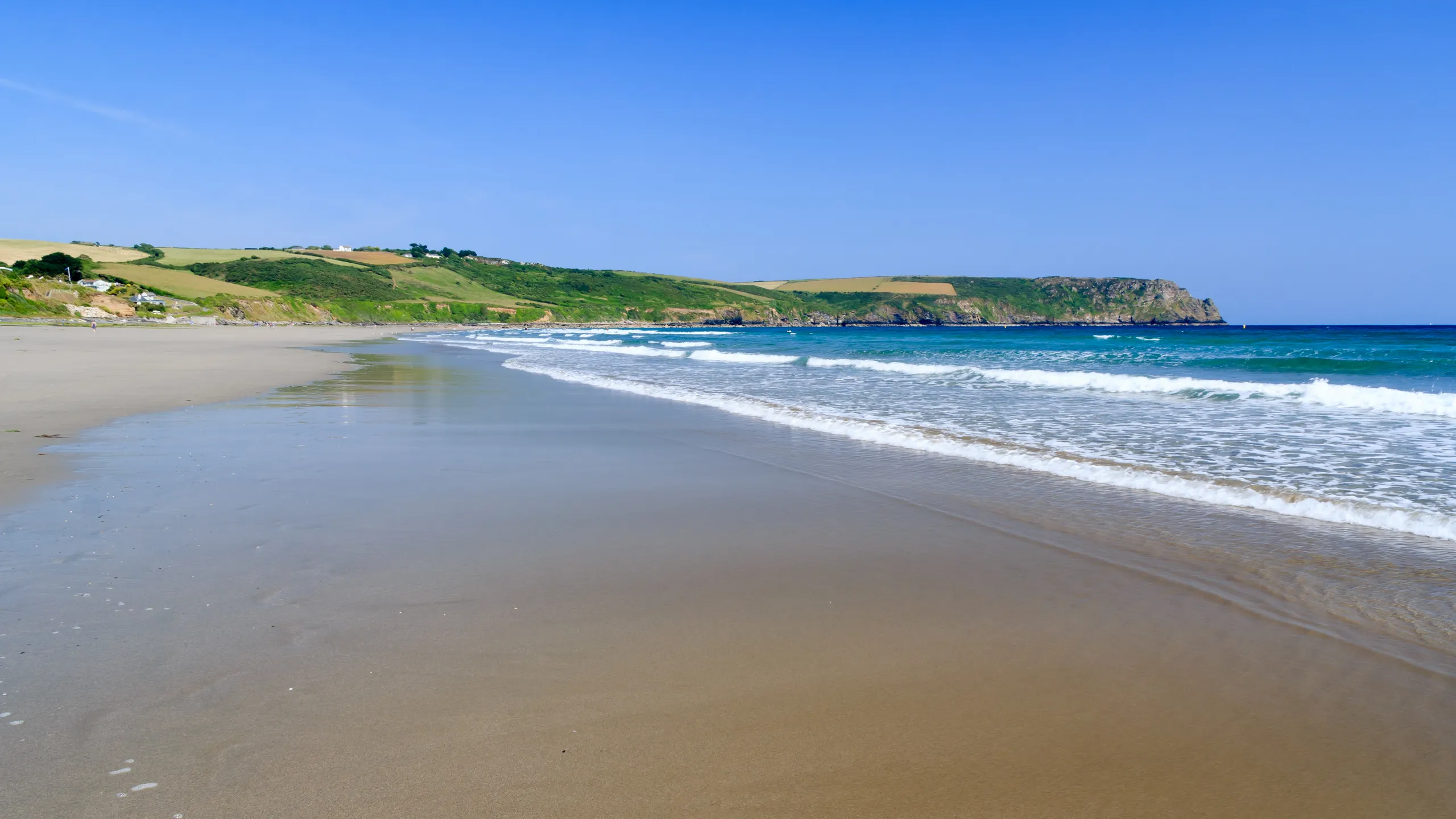 Pendower Beach Cornwall  Wide sandy beach with gentle waves, green hills and a rocky headland beneath a clear blue sky