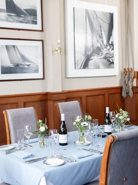 Elegantly set dining table with pale blue linen, floral arrangements and framed sailing photographs on wood-panelled walls