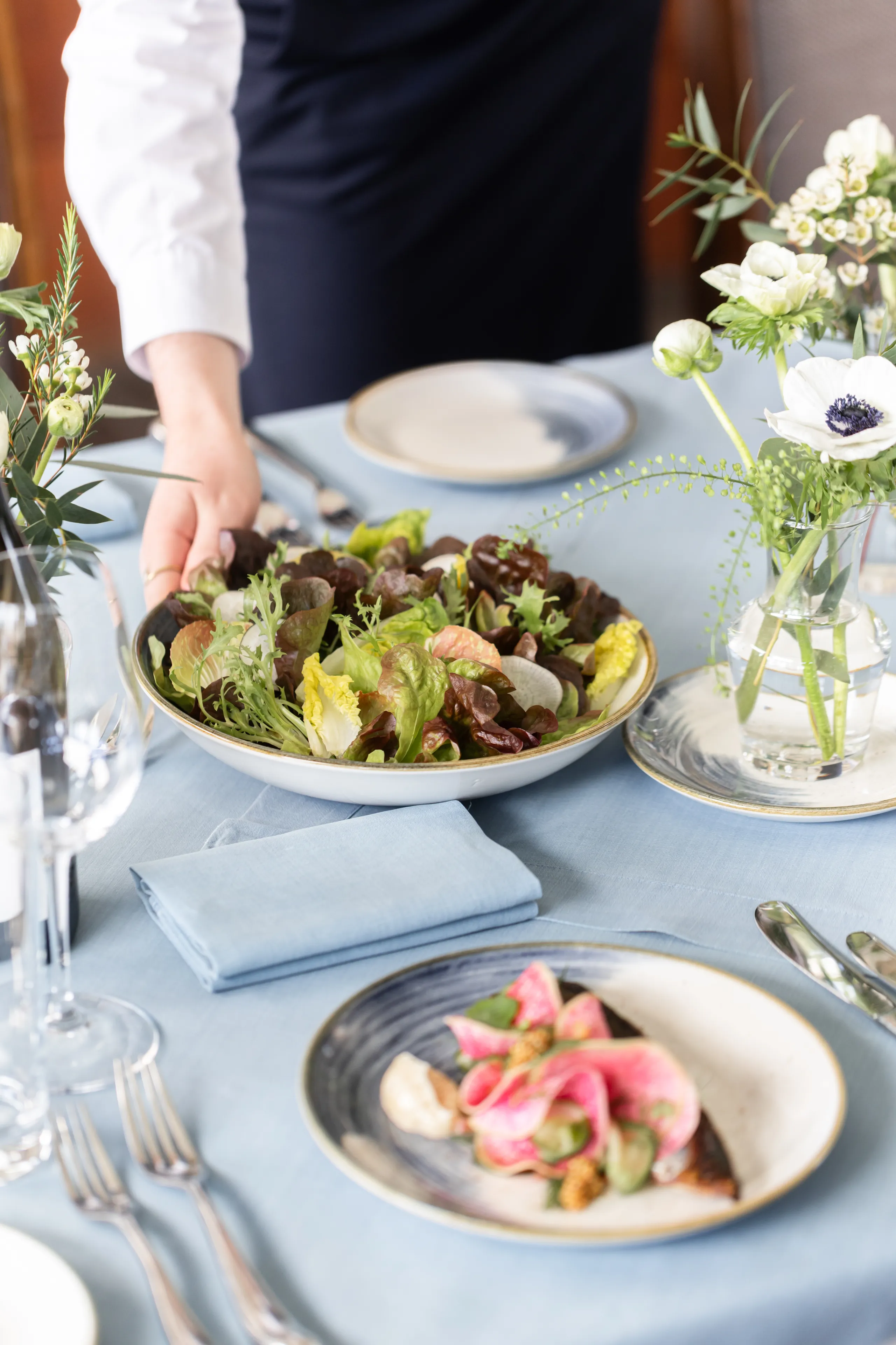 A server places a bowl of mixed leaf salad on an elegantly set dining table with a pale blue tablecloth, floral arrangements and plated food.