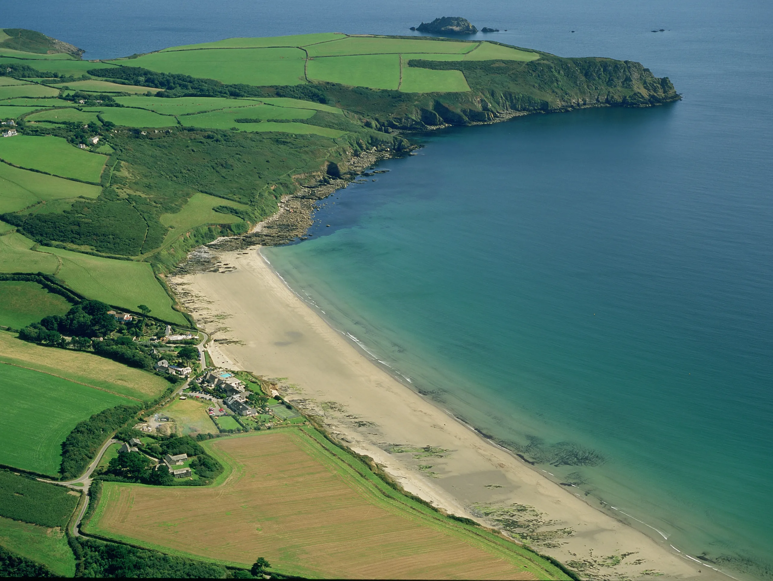 Aerial view of Carne beach beside turquoise water, with patchwork green fields, cliffs at Nare Head and a small coastal hamlet.