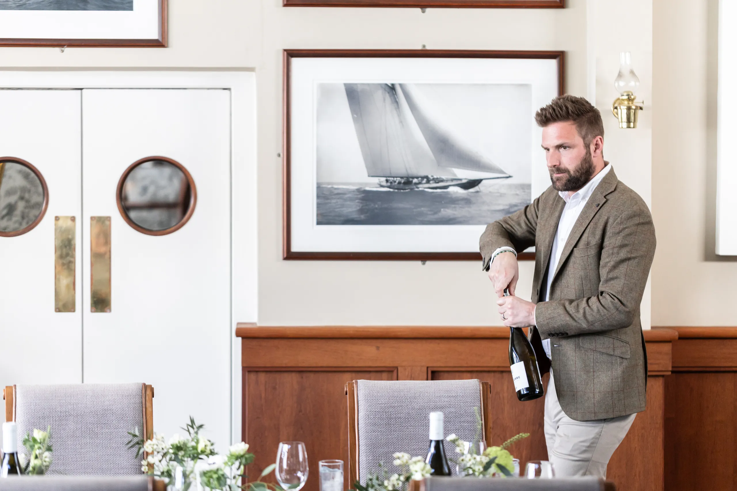 Man opening a bottle of wine in an elegant dining room with framed sailing artwork and laid tables
