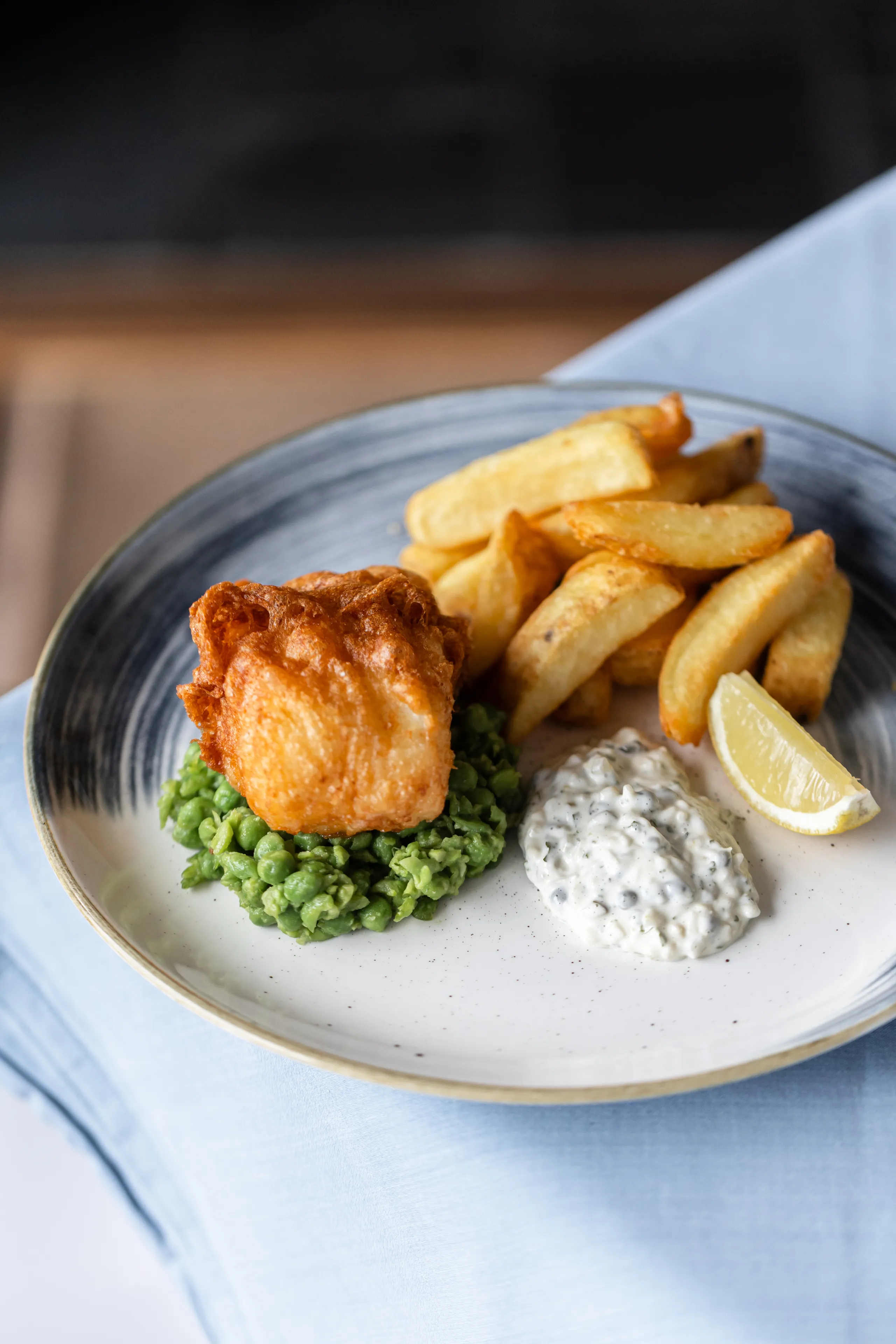 Battered fish with chunky chips, mushy peas, tartare sauce and a lemon wedge on a plate