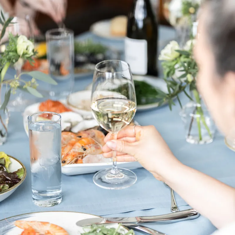 Hand holding a glass of white wine over an elegant seafood dining table with prawns, mussels and floral arrangements