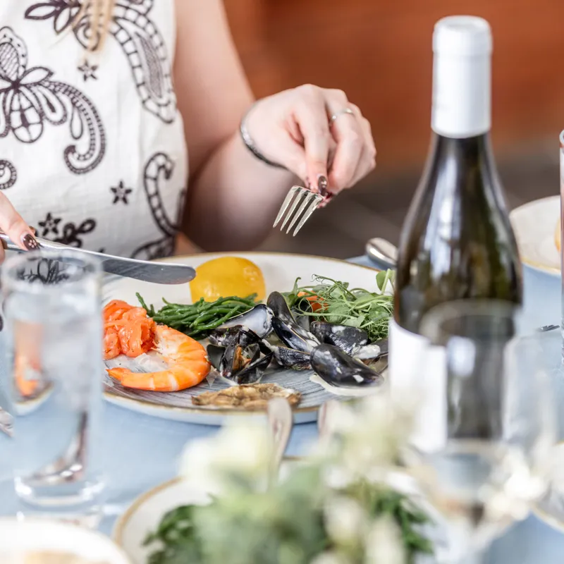 Woman dining on a seafood platter with prawns, mussels and greens at a restaurant table with a bottle of white wine.